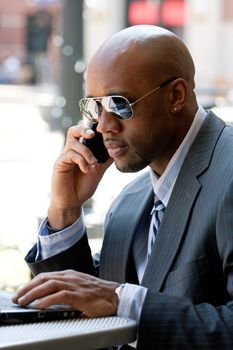 A business man in his early 30s talking on his cell phone and working on his laptop or netbook computer.