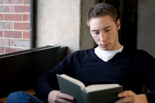 A young male college student reads a book at the library.