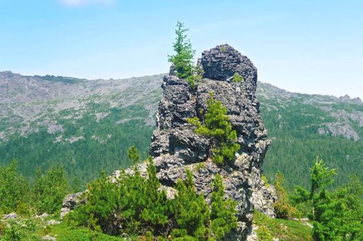 A single rock with trees on top and at the foot against the backdrop of mountains and blue sky