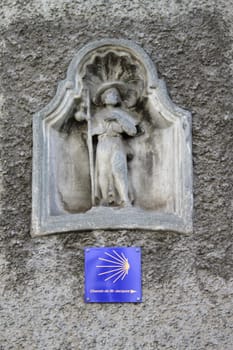 Yellow and blue sign for Compostela way under a pilgrim sculpture on a wall