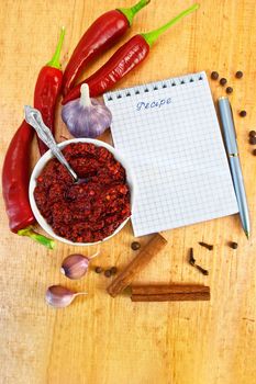 Tabasco in a white cup, fresh red peppers, garlic, cloves, cinnamon, pea black pepper, a notepad with the word recipe and a silver pen on a wooden board