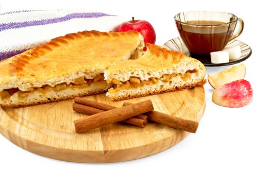 Apple pie with cinnamon sticks on a wooden round board, a whole apple, napkin, tea in the brown cup with two pieces of sugar, two slices of apple isolated on a white background