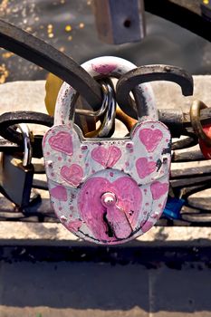 White padlock with painted pink hearts on a close-up on the fence on the background of other castles, water, concrete fences