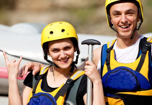 Young happy couple with helmets and rafting equipment, girl showing rock and roll sign