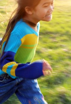 Small girl running happily through green field with speed blur, sidelit by setting sun.