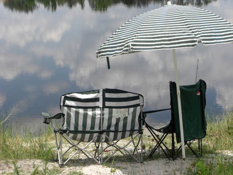 Empty chairs and an umbrella are sitting alongside a lake with a reflection of the sky