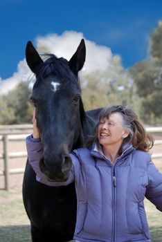 Vertical image of a woman of the Baby Boomer era smiling broadly at her beautiful black horse, against a blue cloudy sky.