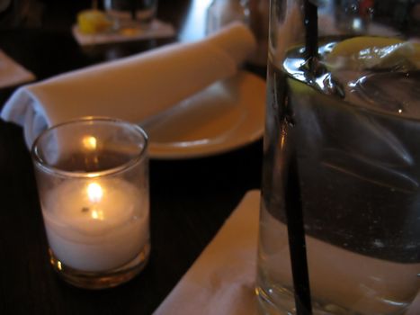 A closeup of a table at a restaurant - waiting to be served.  in the foreground is some water with lemon, and a romantic little candle.