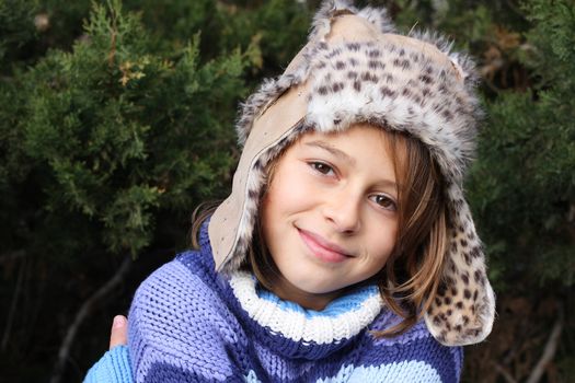 Girl with a fake fur leather hat standing in front of a christmas tree, smiling at the camera. Positive feeling