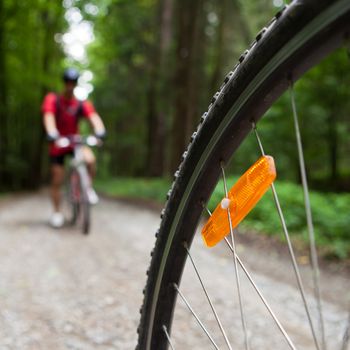 Mountain biking in a forest - bikers on a forest biking trail (shallow DOF, focus on the bike wheel in the foreground)