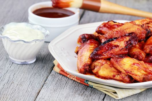 Plate of chicken wings with selectuve focus on chicken at the edge of plate and extreme shallow depth of field. Ranch dressing and BBQ sauce with basting brush in the background. 
