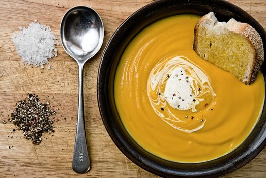 Pumpkin soup in a bowl with toast and cream with a spoon and salt and pepper on the side.