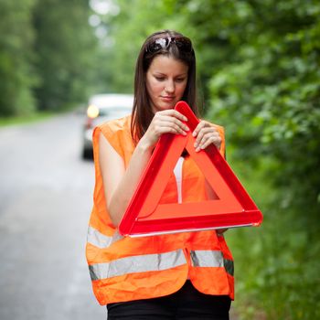 Young female driver wearing a high visibility vest, calling the roadside service/assistance after her car has broken down