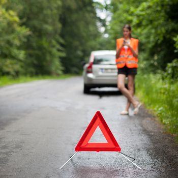 Young female driver wearing a high visibility vest, calling the roadside service/assistance after her car has broken down