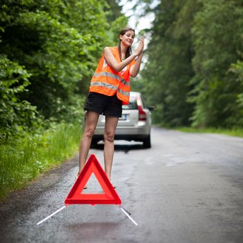 Young female driver wearing a high visibility vest, calling the roadside service/assistance after her car has broken down