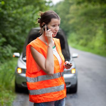 Young female driver wearing a high visibility vest, calling the roadside service/assistance after her car has broken down