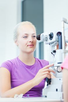 optometry concept - portrait of a young pretty optometrist using slit lamp, checking her patient's sight (color toned image)