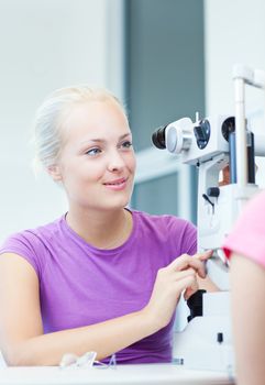 optometry concept - portrait of a young pretty optometrist using slit lamp, checking her patient's sight (color toned image)