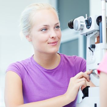 optometry concept - portrait of a young pretty optometrist using slit lamp, checking her patient's sight (color toned image)
