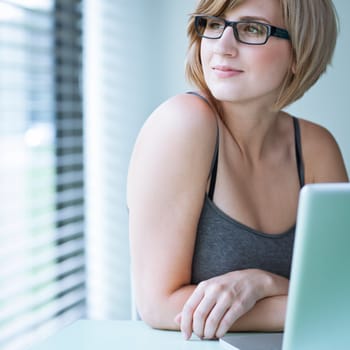 Portrait of a young woman pensively looking out of the window while sitting in front of her laptop