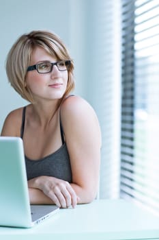 Portrait of a young woman pensively looking out of the window while sitting in front of her laptop