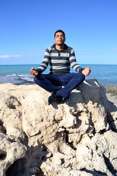 Arab man meditating sitting on rocks at the coast of tunisia.