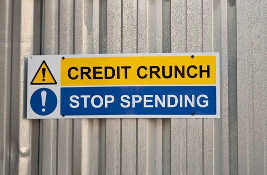 A blue and yellow warning sign with the words CREDIT CRUNCH and STOP SPENDING against metal background