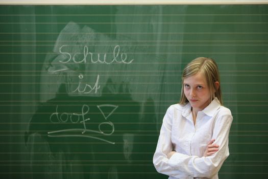Stressed student in the classroom. She stands before a blackboard with the words "school is stupid " and rolls his eyes