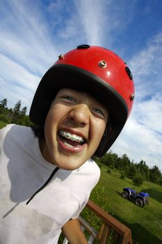 An ecstatic Asian boy with red helmet, getting ready to ride an all terrain vehicle parked in the background.
