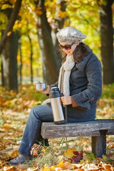 Young pretty woman with glasses and cap drinks tea in autumn park. Vertical view