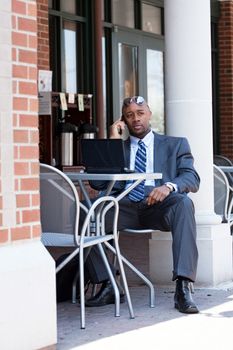 An African American business man in his early 30s talking on his cell phone and using his laptop or netbook computer.