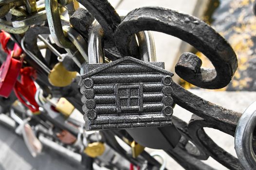 The padlock in the form of house on the iron grating against the background of many other castles, water, concrete pavement