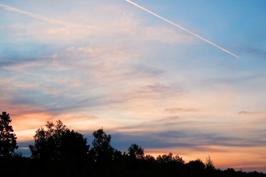 A beautiful sunrise, clouds, painted in red, orange colors, the trees in the meadow, the trail from an airplane in the sky