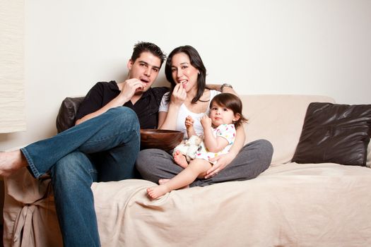Beautiful happy family, father mother toddler daughter child, eating snacks while sitting on sofa couch in livingroom.