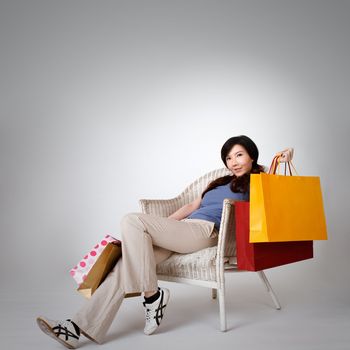 Shopping woman sitting on chair and holding bags, full length portrait isolated on gray studio background.