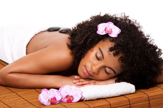 Beautiful happy relaxed Latina-African woman at health day spa with hot stone massage treatment laying in white towel on bamboo table decorated with orchids.