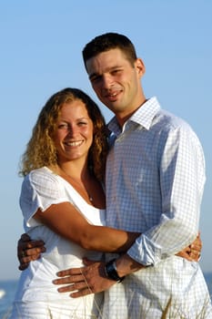 A young happy couple is standing together at the beach with the sea in the background.