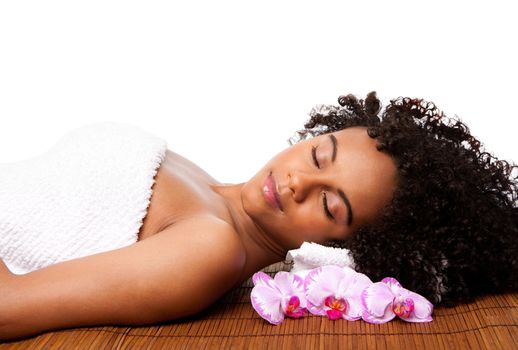 Beautiful happy peaceful sleeping woman at a day spa, laying on bamboo massage table with head on pillow wearing a towel and orchid flowers around, isolated.