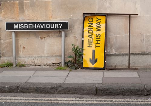 A conceptual scene involving two British street signs situated on a pavement with block wall behind