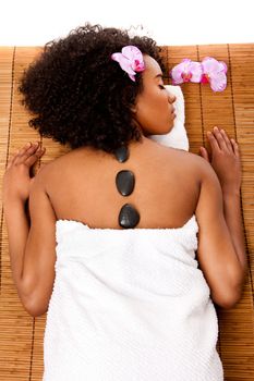 Beautiful happy relaxed Latina-African woman at health day spa with hot lastone therapy massage treatment laying in white towel on bamboo table decorated with orchids.