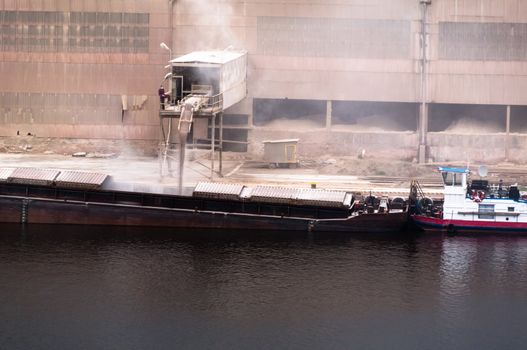 A transport ship is being loaded with powder/grain the oldfashioned way with dust all over