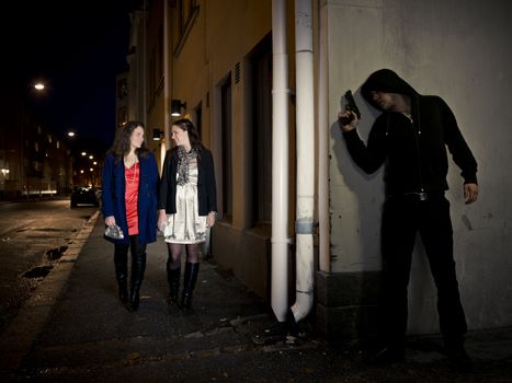 Hooded man stalking two women behind a corner holding a gun
