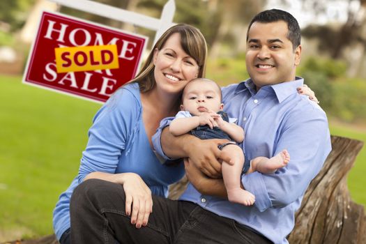 Happy Mixed Race Couple with Baby in Front of Sold Real Estate Sign.