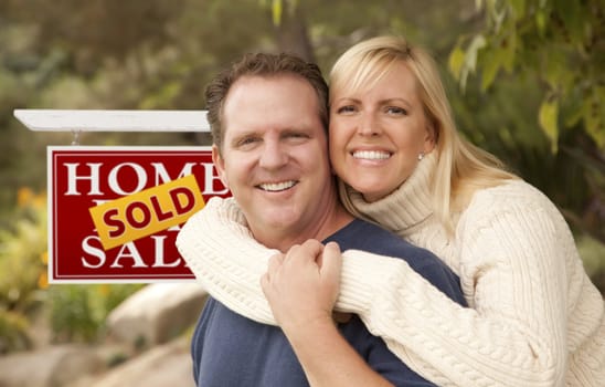 Happy Attractive Caucasian Couple in Front of Sold Real Estate Sign.