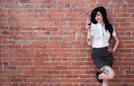 Tattooed girl with a glass of red wine leaning against a brick wall.