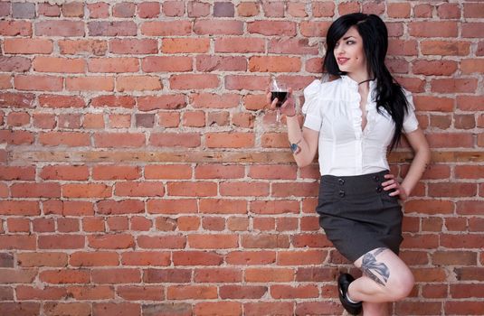 Black-haired tattooed girl with a glass of red wine leaning against a brick wall.