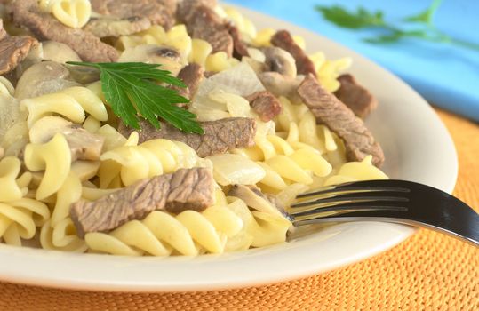 Veal strips with white mushrooms and onion in cream sauce served on fusilli pasta and garnished with a parsley leaf (Selective Focus, Focus on the parsley, the meat below and the front of the fork)