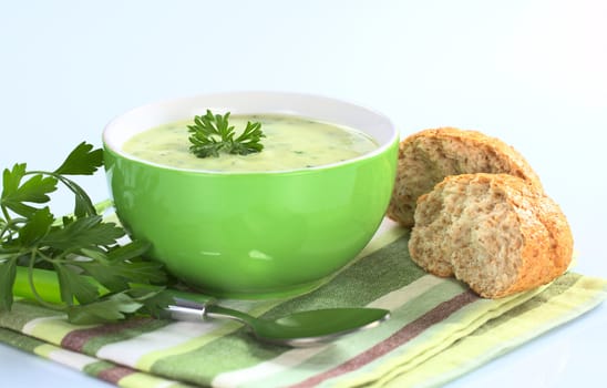 Cream of potato with herbs and green onions garnished with parsley and served in a green bowl with parsley and buns on the side photographed on bluish background (Selective Focus, Focus on the front of the parsley in the soup)