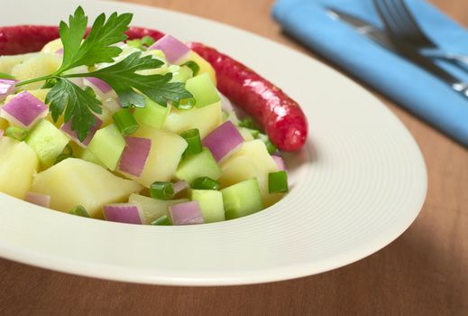 Potato salad with cucumber, red and green onions with oil dressing garnished with parsley and served with small sausages in the back (Selective Focus, Focus on the parsley and the salad around)