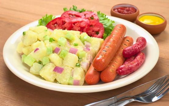 Sausages and potato salad with red and green onions and cucumber with a mayonnaise-cream dressing and tomato slices on plate with ketchup and mustard in the back (Selective Focus, Focus on the front of the meal)